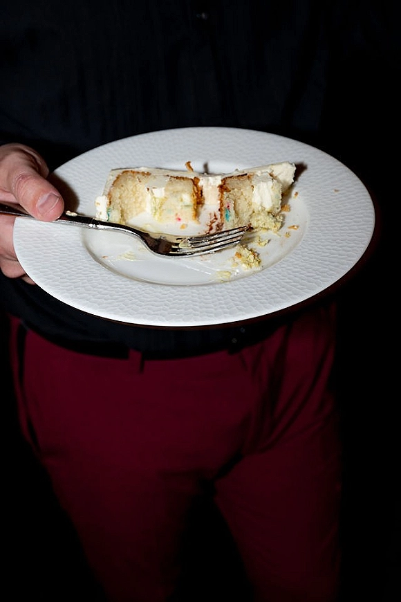 Wedding cake slice on a white dessert plate with fork and buttercream frosting, held by a person in a dark shirt and pants