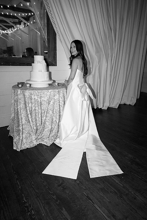 Bride portrait beside a tiered white wedding cake, wearing a strapless gown with oversized bow and long train under string lights indoors