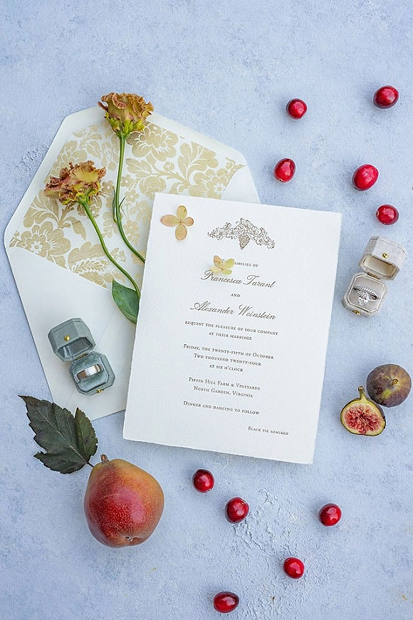 Wedding invitation suite flat lay with invitation card, patterned liner, rings in boxes, flowers and fig, pear, red berries on light blue surface