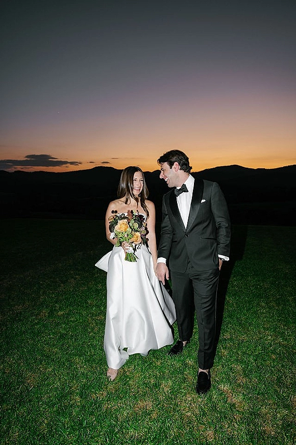 Couple portrait of bride and groom laughing as they walk at sunset, bride holding a bouquet against mountains and a grassy field
