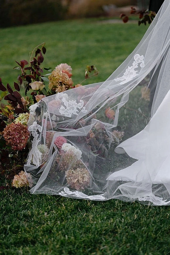 Bridal veil detail with lace edge veil draped over roses and hydrangeas, arranged on a grass lawn with greenery and burgundy foliage