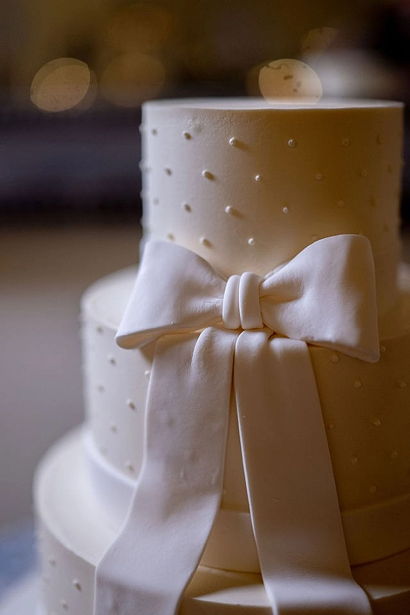 Wedding cake with smooth white frosting and a fondant bow, ribbon drape, and pearl dot piping, set indoors with blurred lights