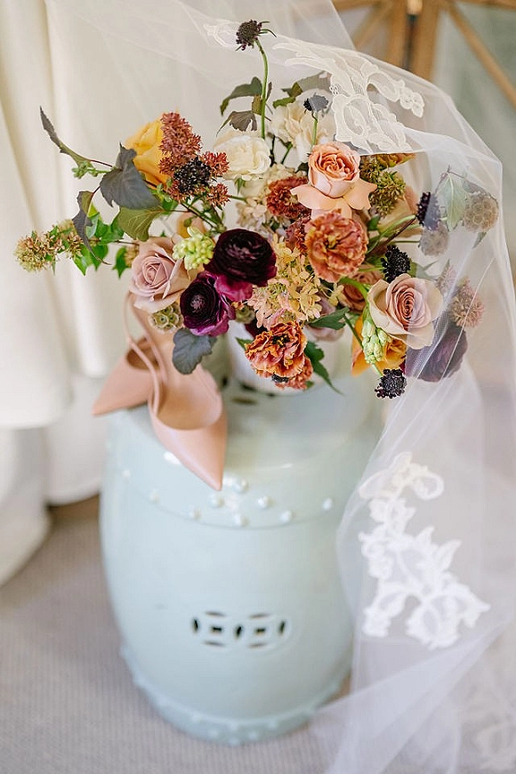 Bridal bouquet styled with lace veil and pointed toe heels on a pastel pedestal, with scattered pearls against white curtains