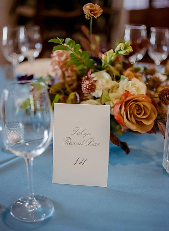 Wedding place card with calligraphy beside a rose centerpiece on a blue tablecloth, surrounded by wine glasses on a dim reception table