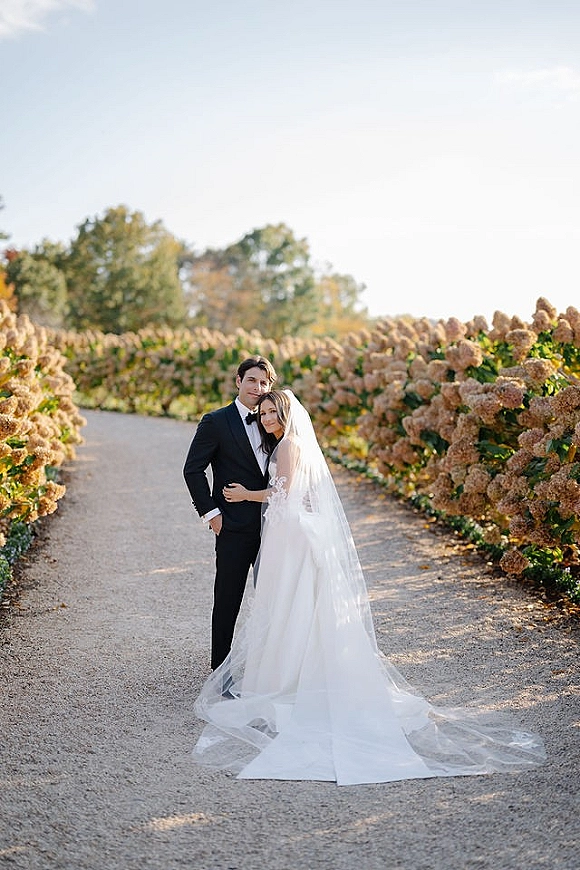 Couple portrait of bride and groom embrace on a garden path, her long veil and lace dress train flowing beside hydrangeas
