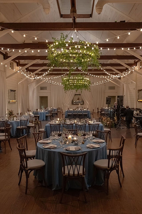 Reception tablescape with round wedding tables in blue linens, candles and low floral centerpieces beneath a greenery chandelier in a white barn