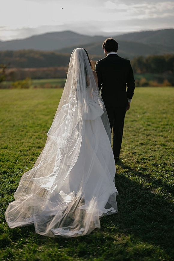 Wedding couple portrait of bride and groom walking away, her long cathedral veil trailing over a grassy field with mountains behind