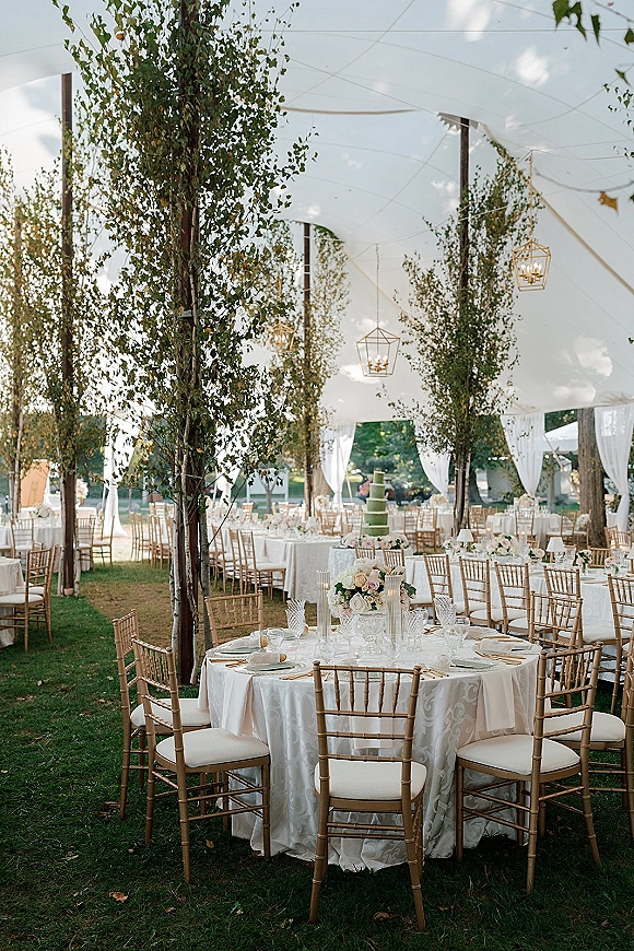 Reception tablescape at an outdoor tented reception with white linens, floral centerpieces, taper candles, and gold chiavari chairs on a garden lawn