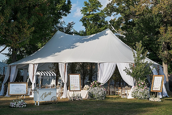 Wedding tent decor with white draped tent entrance, framed welcome and seating chart signs, lanterns, and gold chiavari chairs on a lawn