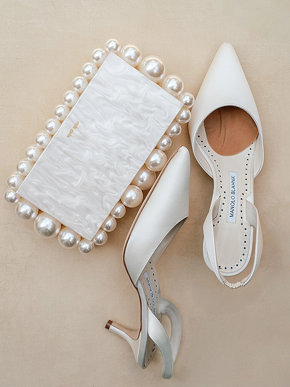 Bridal shoes and white wedding heels in a flat lay with a pearl-embellished clutch on a neutral beige surface