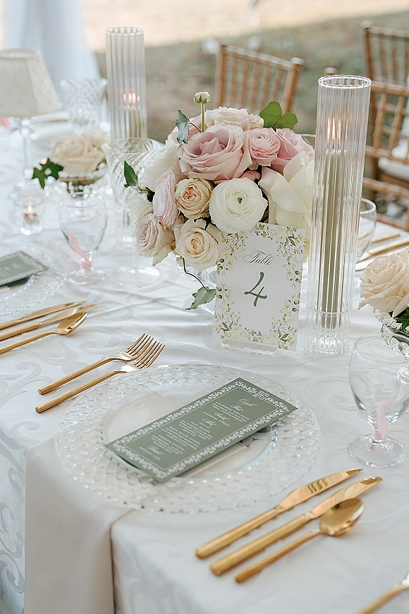Reception tablescape with a wedding table centerpiece of blush and ivory roses, ribbed glass candle holders, and gold flatware on a lawn setting