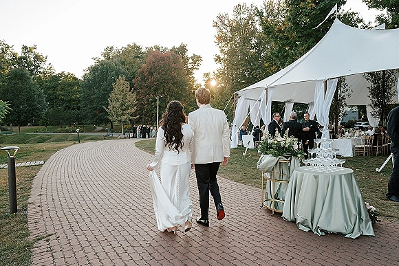 Newlywed walk as bride and groom stroll away on a brick pathway toward a white tent reception, bride lifting her train in sunset light