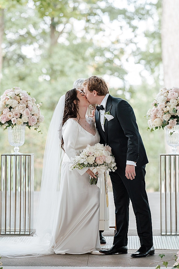 Wedding kiss as bride in long sleeve gown and veil holds blush and white rose bouquet, groom in tuxedo on terrace with columns and greenery