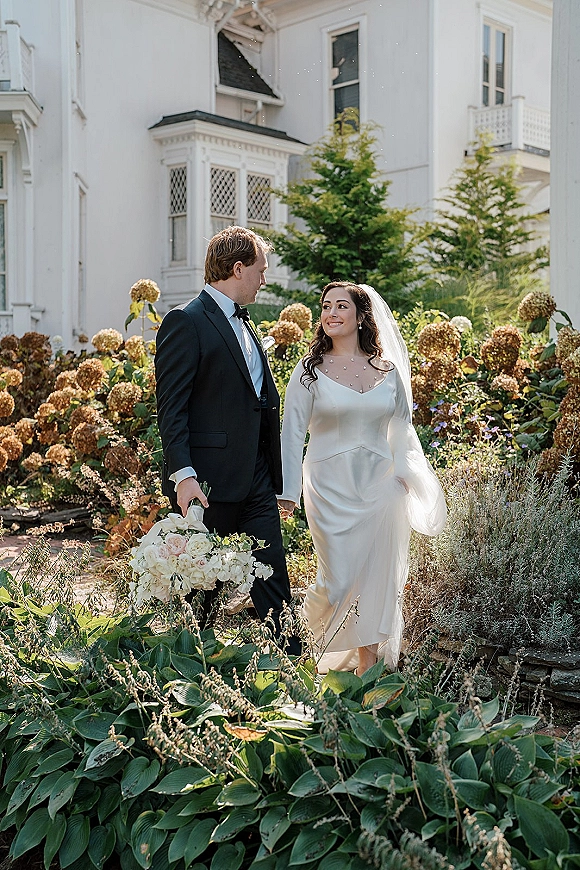 Couple portrait of bride and groom walking hand in hand, her veil and white bouquet in sunlight by hydrangeas and a white estate house