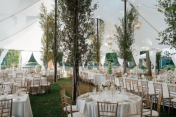 Reception tent decor with white draped tent accents, greenery-wrapped poles, hanging lanterns, round tables, and tiered cake on a lawn