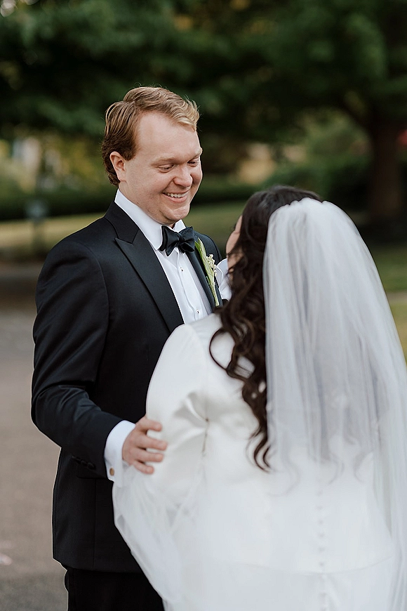 Wedding couple portrait of bride and groom smiling, holding hands in a garden path, with a flowing bridal veil accent and tuxedo bow tie