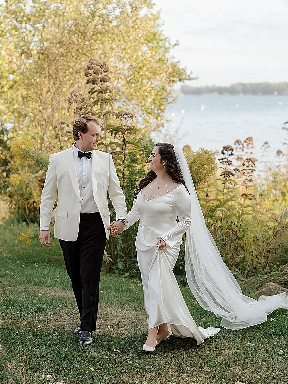 Couple portrait of bride and groom holding hands, walking by a lakeside with greenery as her long veil trails behind them.