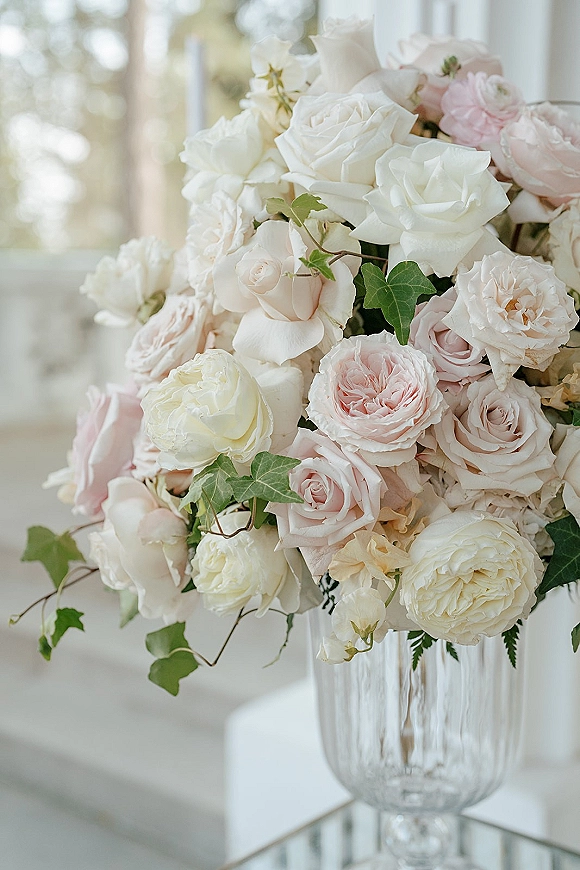 Wedding centerpiece with blush and white roses in a glass vase, accented with ivy greenery, lit by soft window light in a white room