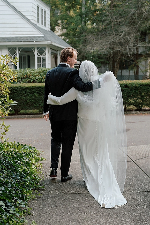 Couple portrait of bride and groom walking away arm in arm, her cathedral veil trailing over a button-back gown by garden shrubs and house exterior