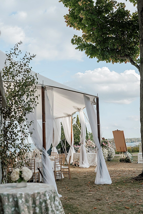 Outdoor reception decor under wedding tent draping with round tables, lace cloths, gold Chiavari chairs, and floral centerpieces on a lawn near water