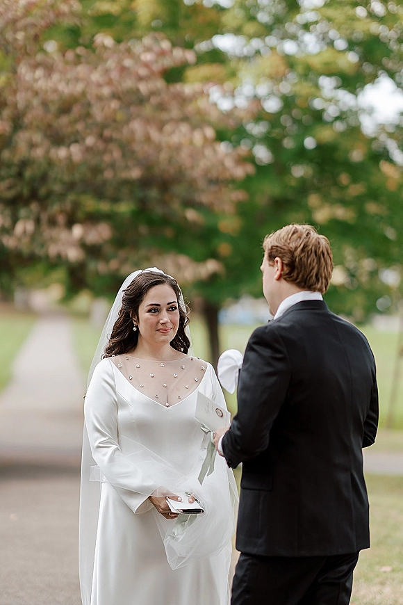 Wedding vows as groom reads from a vow book to bride in long sleeve pearl neckline gown and veil on a tree-lined park path