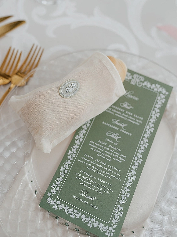 Wedding menu card in green with floral border on a white napkin sealed with wax, set on a clear charger with gold flatware