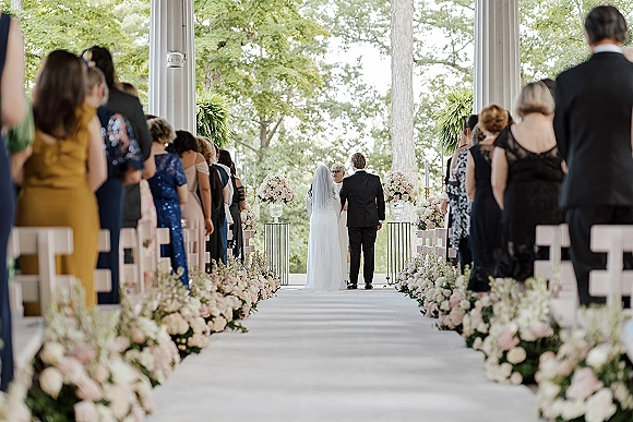 Ceremony moment as bride and groom stand at the altar during wedding vows, seen from behind with a long veil and floral-lined aisle in an outdoor pavilion