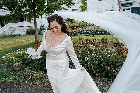 Bridal portrait of a bride with veil in a long sleeve wedding dress, holding an invitation suite on a garden pathway by a white house porch