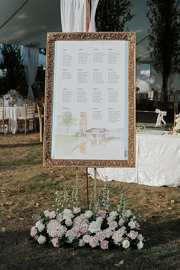 Wedding seating chart in an ornate gold frame on a stand, with roses and hydrangeas at the base under a draped outdoor tent