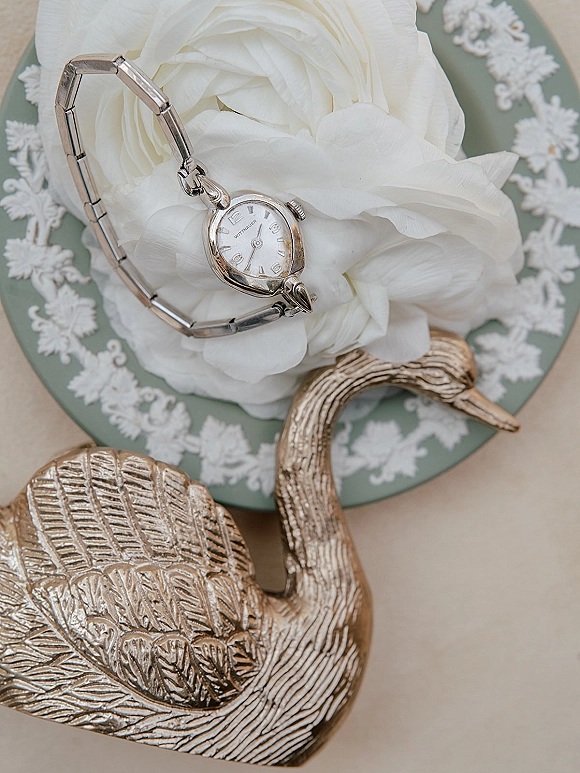 Wedding watch detail flat lay with a groom watch flat lay, white flower and gold swan figurine on lace doily beside a green patterned plate