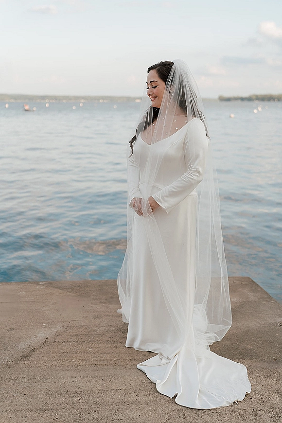 Bridal portrait of a bride in a long sleeve wedding dress with a wedding veil over her face on a concrete pier by the ocean shore