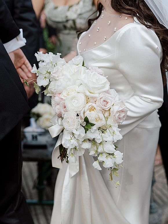 Bridal bouquet with cascading bridal bouquet of white and blush roses, greenery and satin ribbon tails held by bride in pearl veil indoors