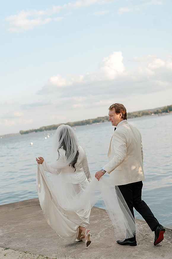 Couple portrait of bride and groom walking on a concrete dock by a lake, long veil blowing in wind under cloudy sky