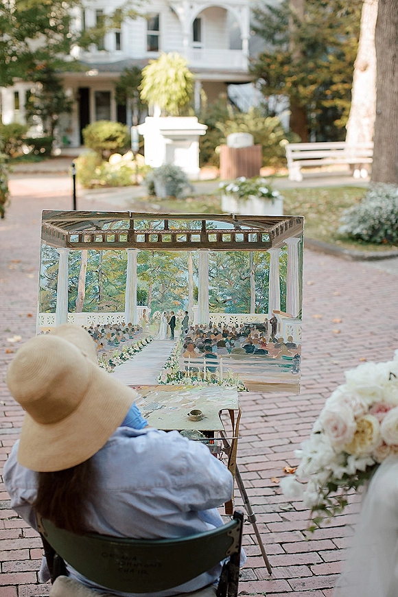 Live wedding painting setup with easel and canvas, paint palette and brushes on a brick walkway near garden trees and a white porch