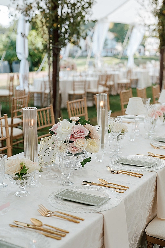 Reception tablescape with an outdoor wedding tablescape featuring blush rose centerpiece, glass hurricane candles, and gold flatware under a draped canopy