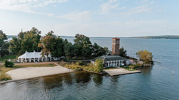 Waterfront wedding venue with a white event tent and lounge chairs on the lawn, overlooking a lake, pier, trees, and clock tower building