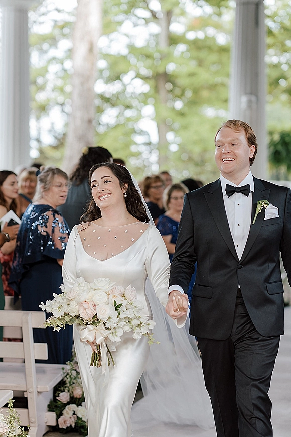 Wedding recessional as bride and groom walk hand in hand down the aisle, bride holding a white bouquet under pavilion columns with guests watching