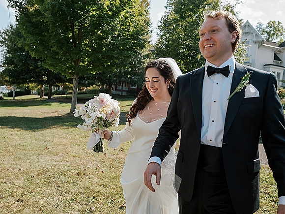 Bride and groom portrait of newlyweds walking on a sunlit lawn, bride holding a bouquet and veil flowing beside groom in black tuxedo