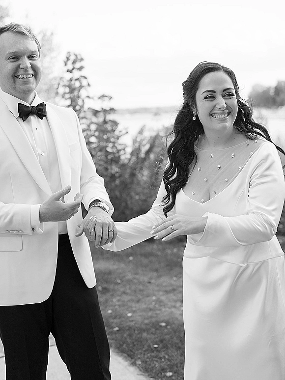 Couple portrait in a black and white wedding photo, bride and groom laughing and holding hands on a lawn by water and trees