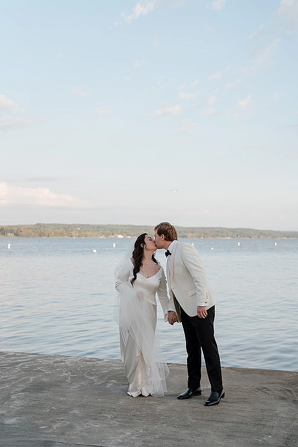 Wedding kiss portrait of bride and groom kissing and holding hands on a concrete pier by a lake, her veil flowing in the breeze