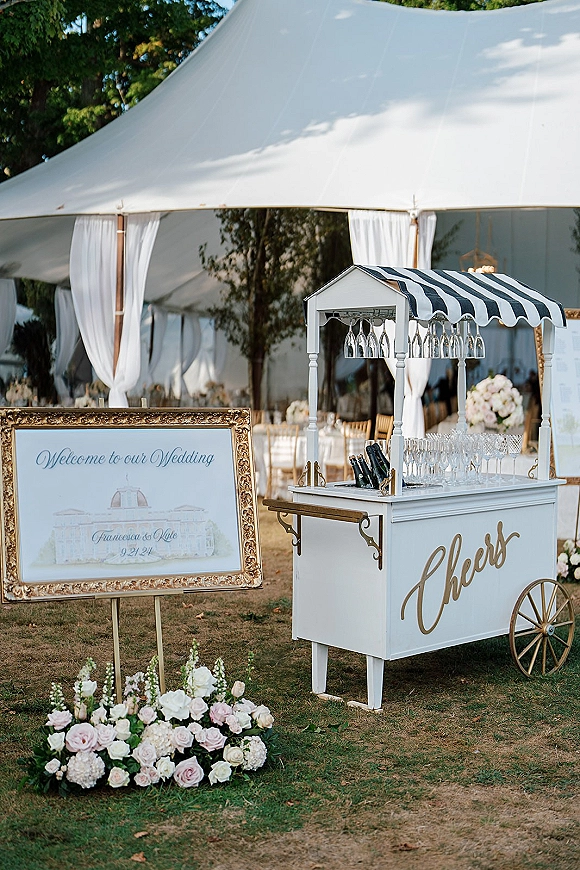 Wedding champagne cart with striped canopy, champagne bottles and coupes, hanging glasses, and gold welcome sign under a draped tent