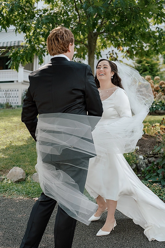 Wedding first look as bride in long sleeve dress laughs with groom in black tuxedo, her veil blowing on a garden lawn by trees