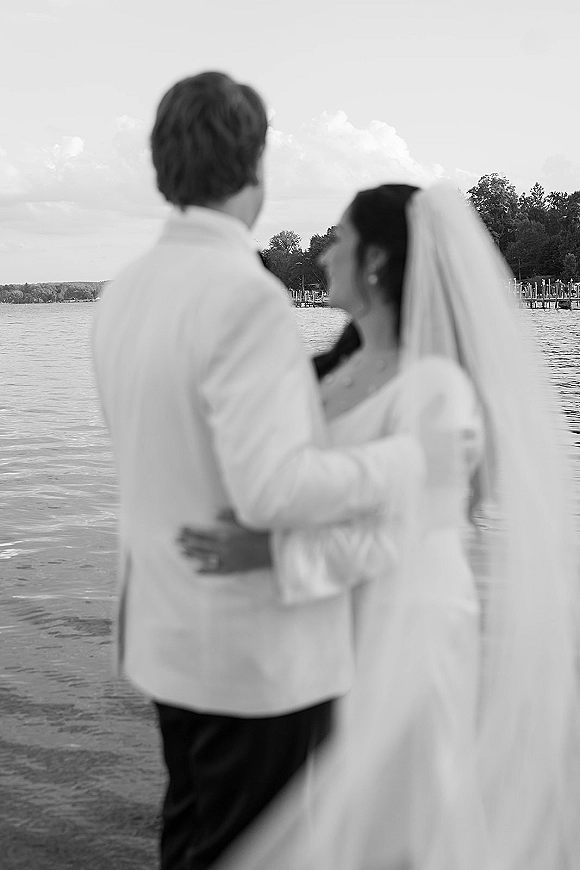 Couple portrait in a black and white wedding photo, bride in veil and groom in suit jacket embracing by a lakeside dock with trees and clouds