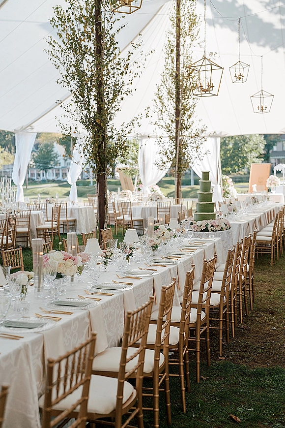 Wedding reception tablescape in an outdoor tent reception with white linens, gold chiavari chairs, floral centerpieces, and candlelit place settings under chandeliers
