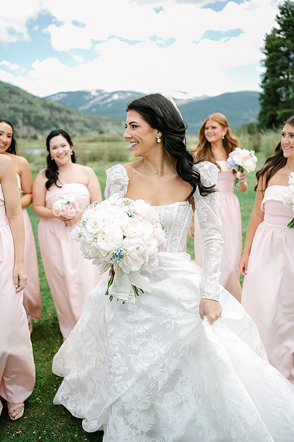 Bridal party portrait of bride with bridesmaids in blush dresses, bride holding bouquet and veil on a grass lawn with mountains behind