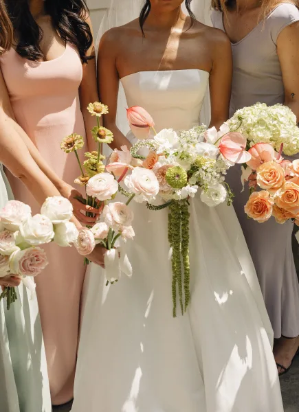 Bridesmaid bouquets of peach and white blooms with roses, ranunculus and anthurium, ribbon-wrapped stems, against a sunlit white wall