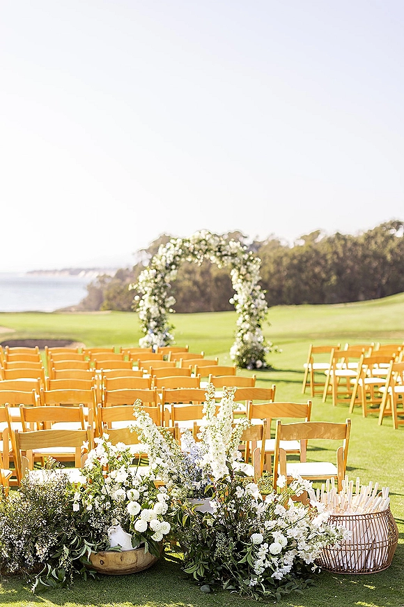 Ceremony setup with wooden folding chairs facing a floral arch of white flowers and greenery, with aisle candles on a lawn overlooking the ocean