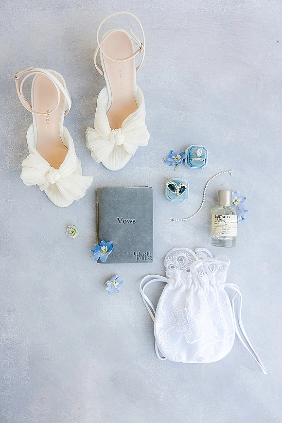 Bridal accessories flat lay featuring white bow heels, vow book, ring boxes and engagement ring, pearl bracelet, perfume, and blue flowers on gray surface