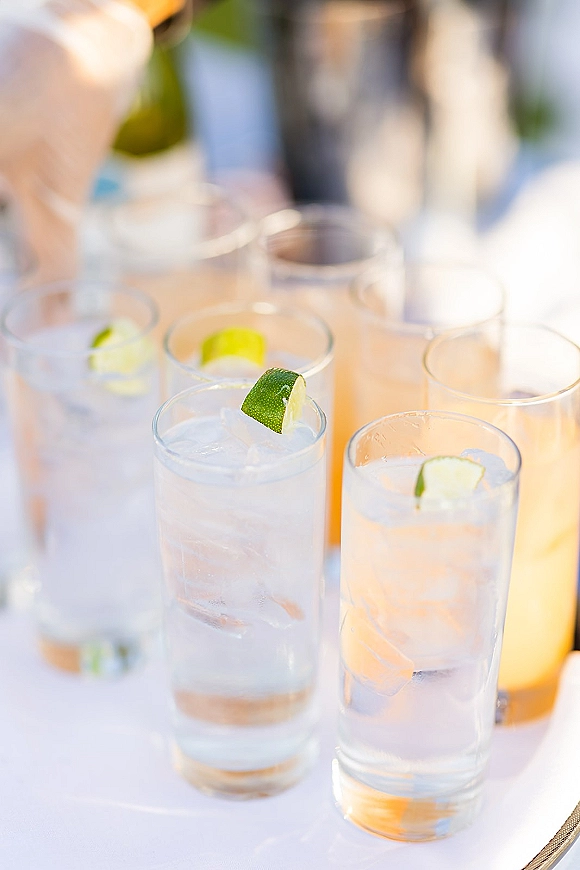 Wedding signature drinks in highball glasses with ice and lime wedges on a white tablecloth at an outdoor bar setup in soft light