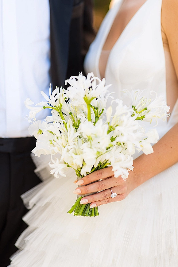 Bridal bouquet of white flowers with long green stems, held by bride with oval engagement ring and nude manicure, groom blurred behind